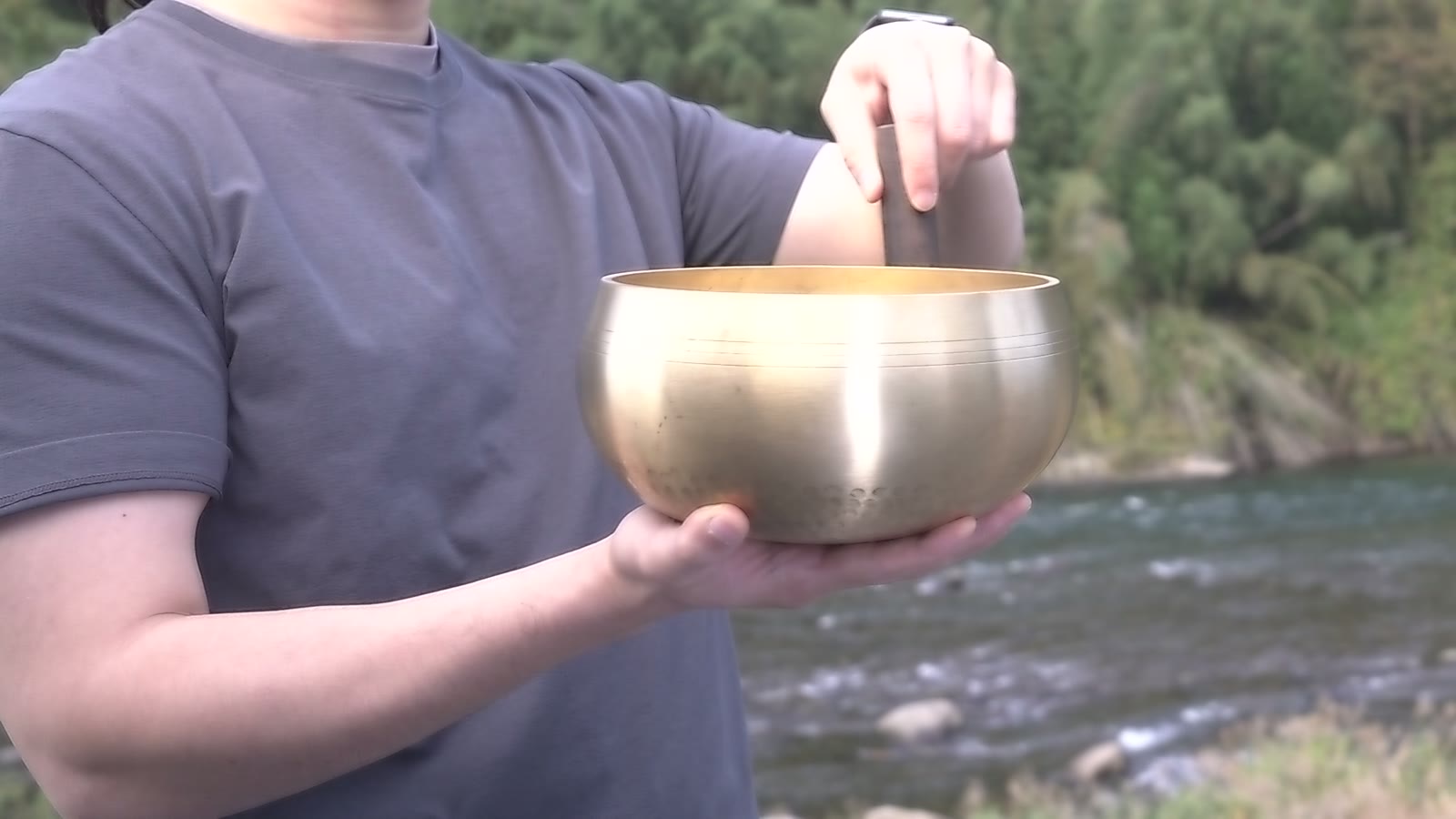 Hands rubbing a singing bowl by a river